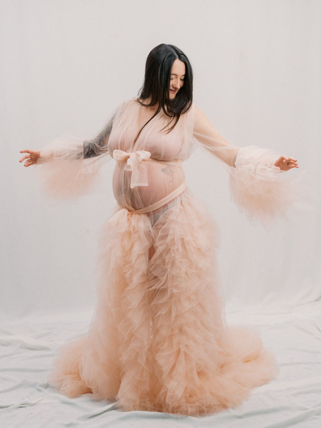 Pregnant woman in a flowing peach tulle maternity gown with bell sleeves and bow, posing in a bright studio.