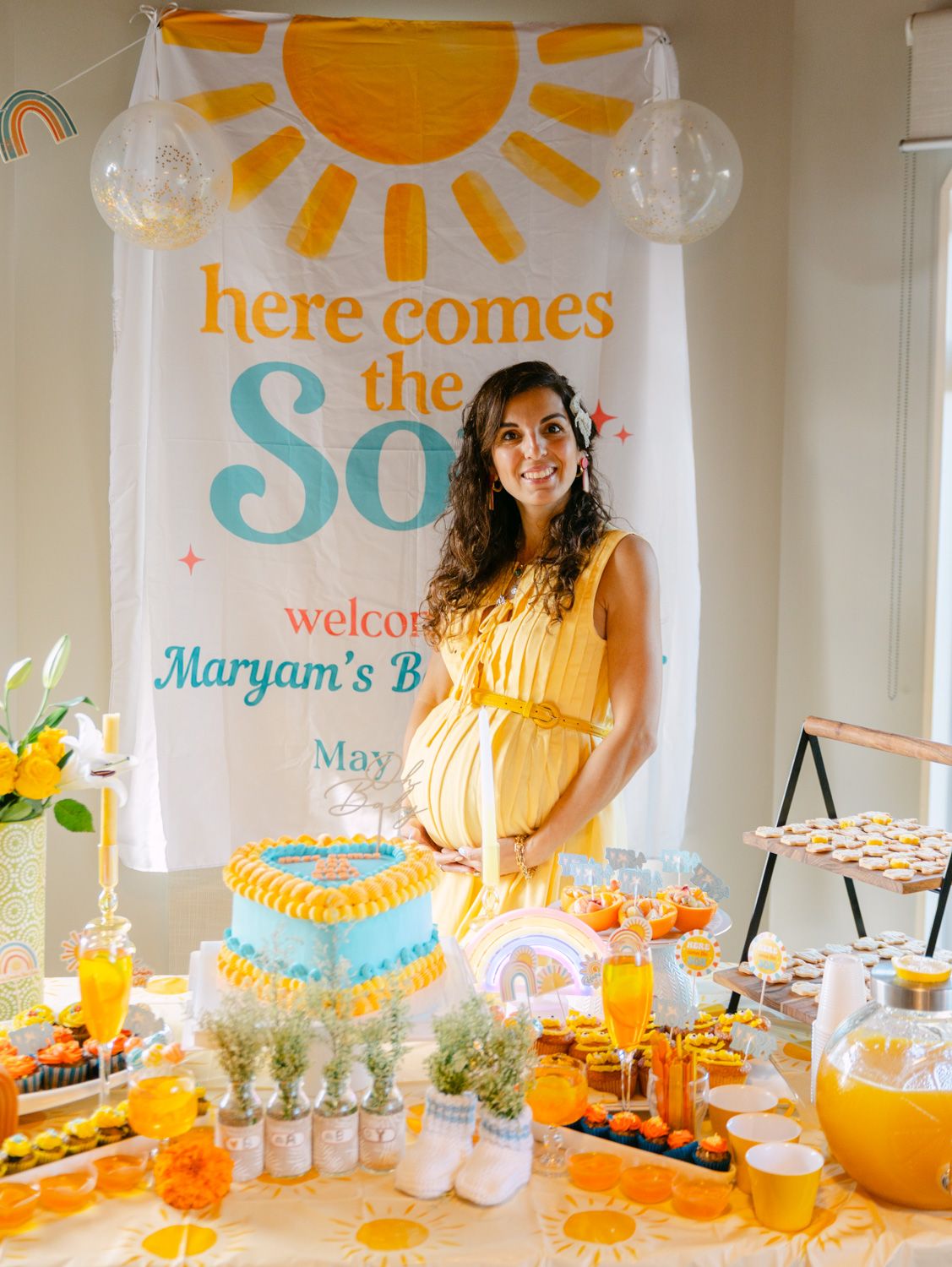 Pregnant woman in yellow dress at a sunshine-themed baby shower with decorated dessert table and Here Comes the Son banner.