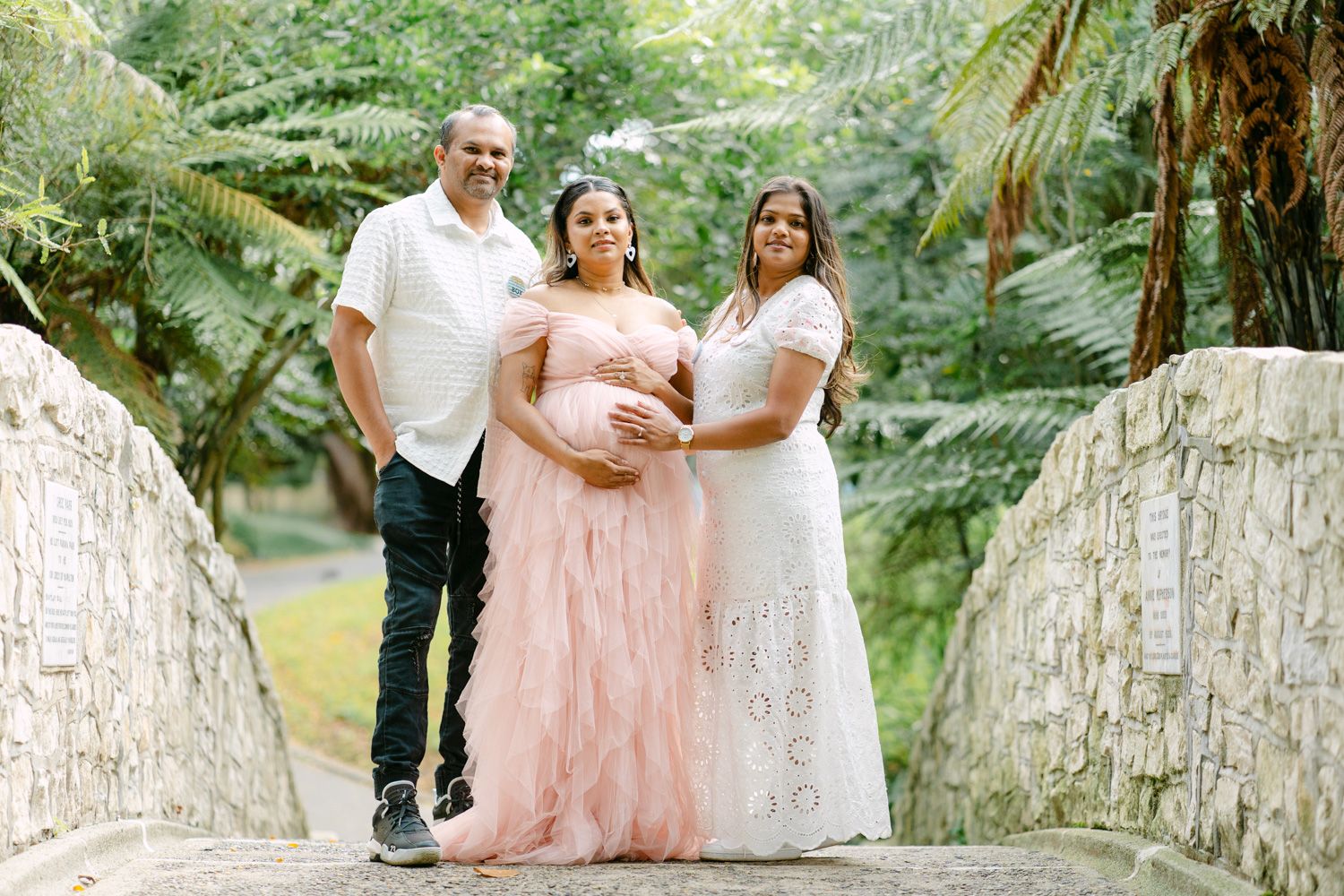 Pregnant woman in pink gown poses with two loved ones on a stone bridge surrounded by lush greenery.
