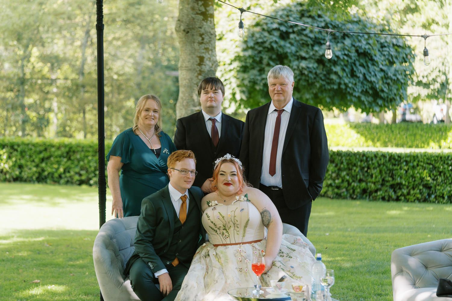 Wedding party of five posing outdoors on green lawn, bride in gold gown seated with groom, three guests standing behind.