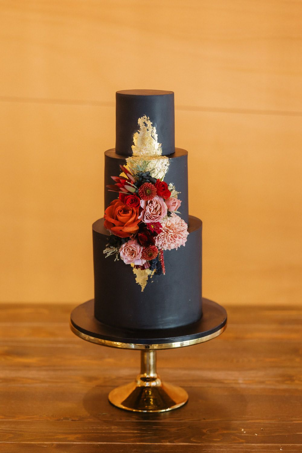 Elegant three-tier black wedding cake with red, pink roses and gold accents on a gold stand.