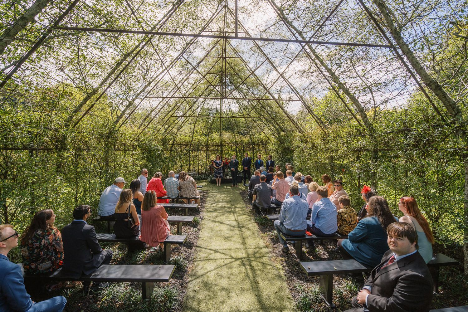 Outdoor wedding ceremony in a living willow structure with guests seated on wooden benches surrounded by lush greenery.