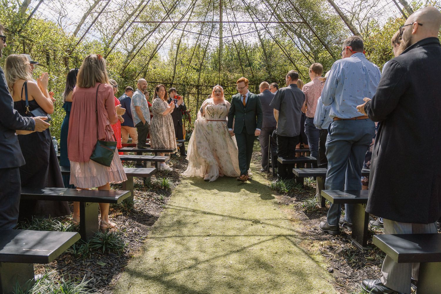 Newlyweds walk down aisle through garden ceremony venue surrounded by guests after wedding.