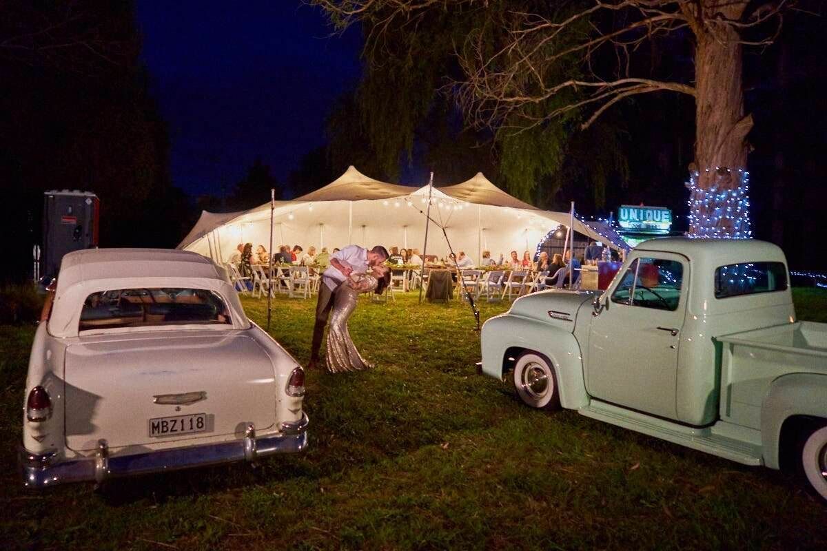 Vintage white cars parked outside a glowing wedding tent at night with fairy lights and dancing guests.