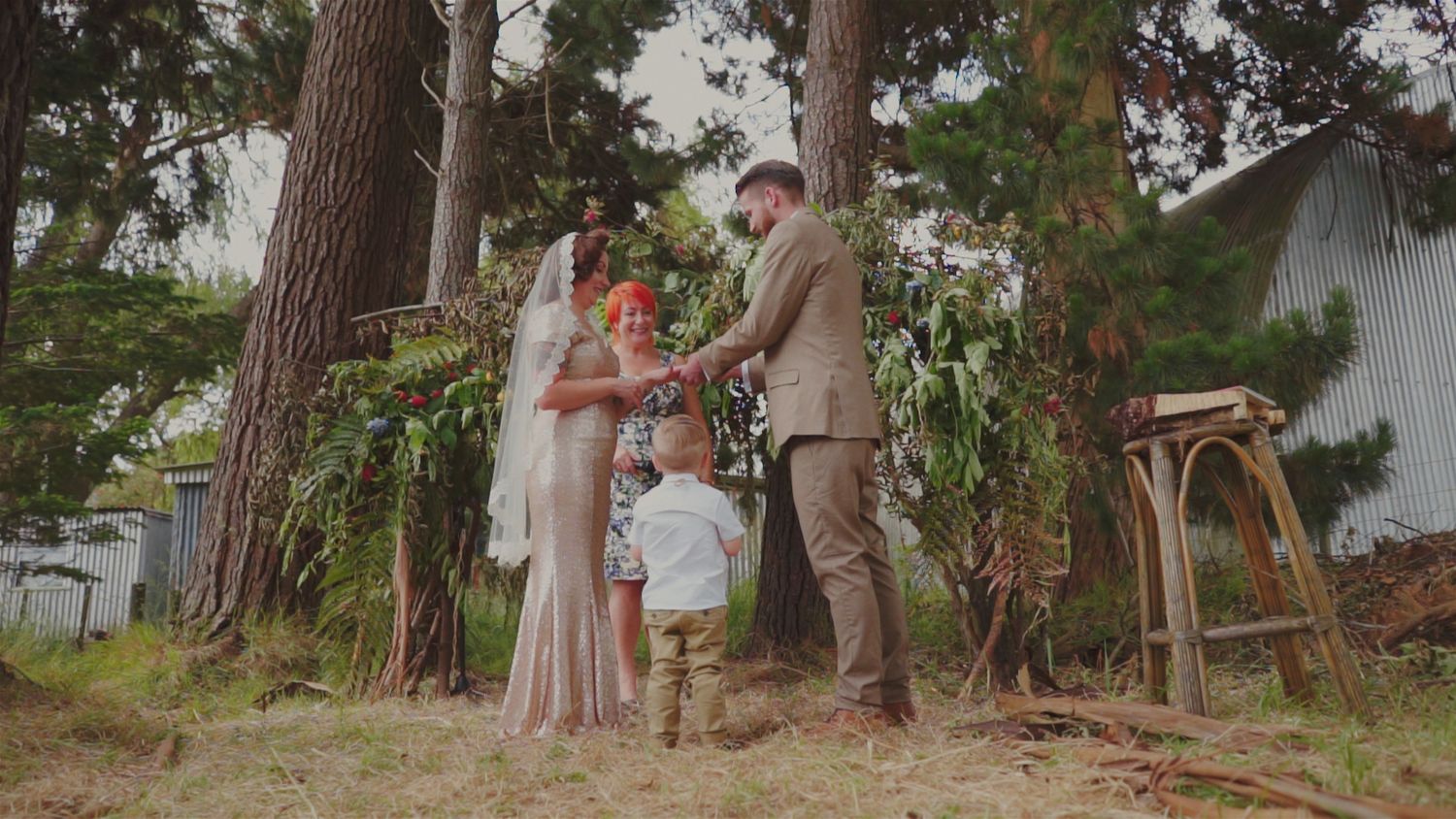 Couple exchanging vows outdoors under tall pine trees with a child present at an intimate woodland wedding ceremony.