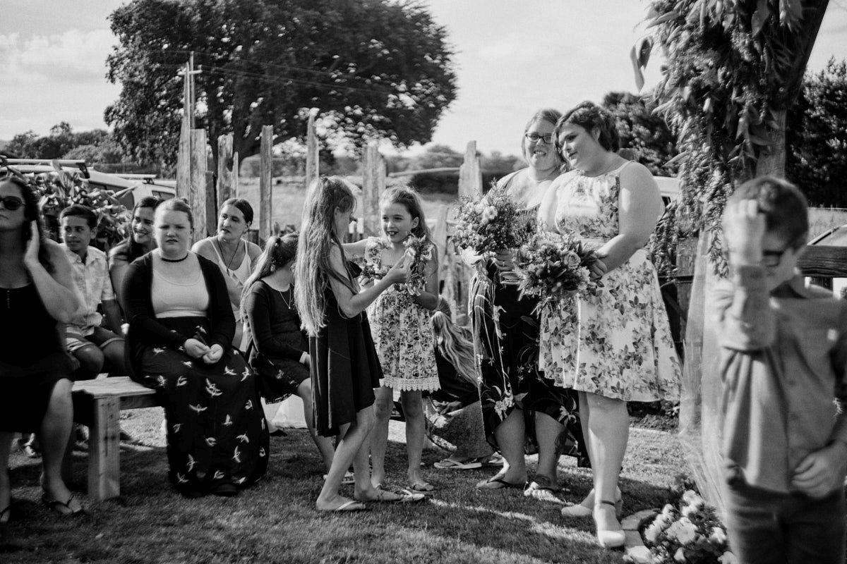 Black and white outdoor wedding ceremony with bridesmaids and flower girls exchanging bouquets among seated guests.