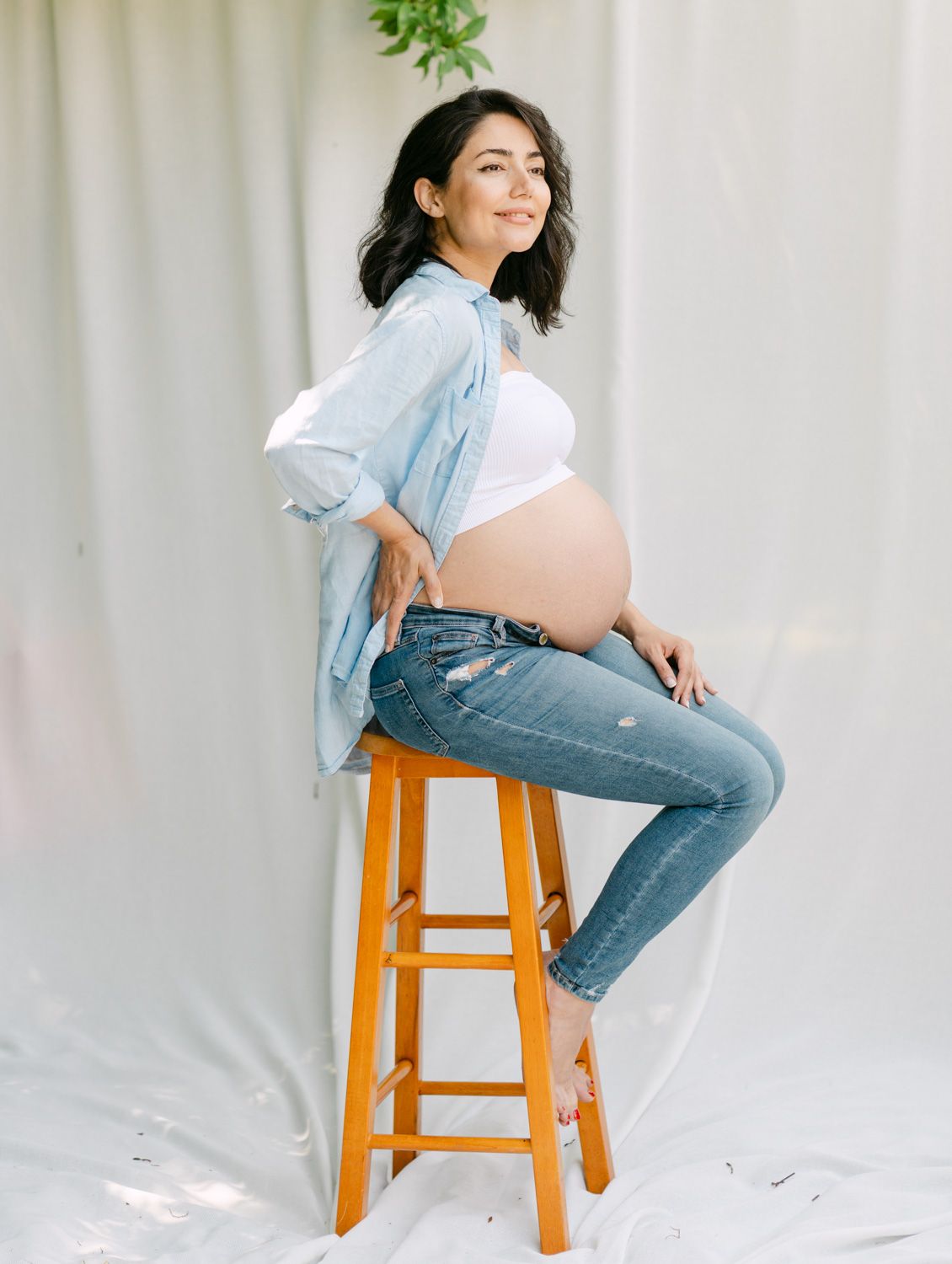 Smiling pregnant woman sitting on wooden stool wearing blue shirt, white crop top, and jeans, showing baby bump.