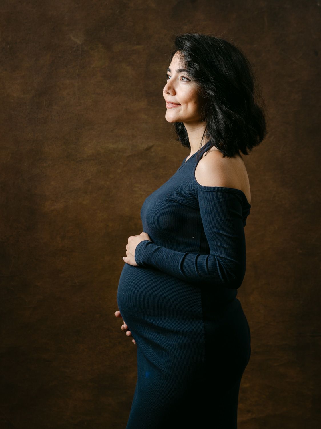 Pregnant woman in navy blue off-shoulder dress cradling baby bump, smiling sideways against brown studio backdrop.