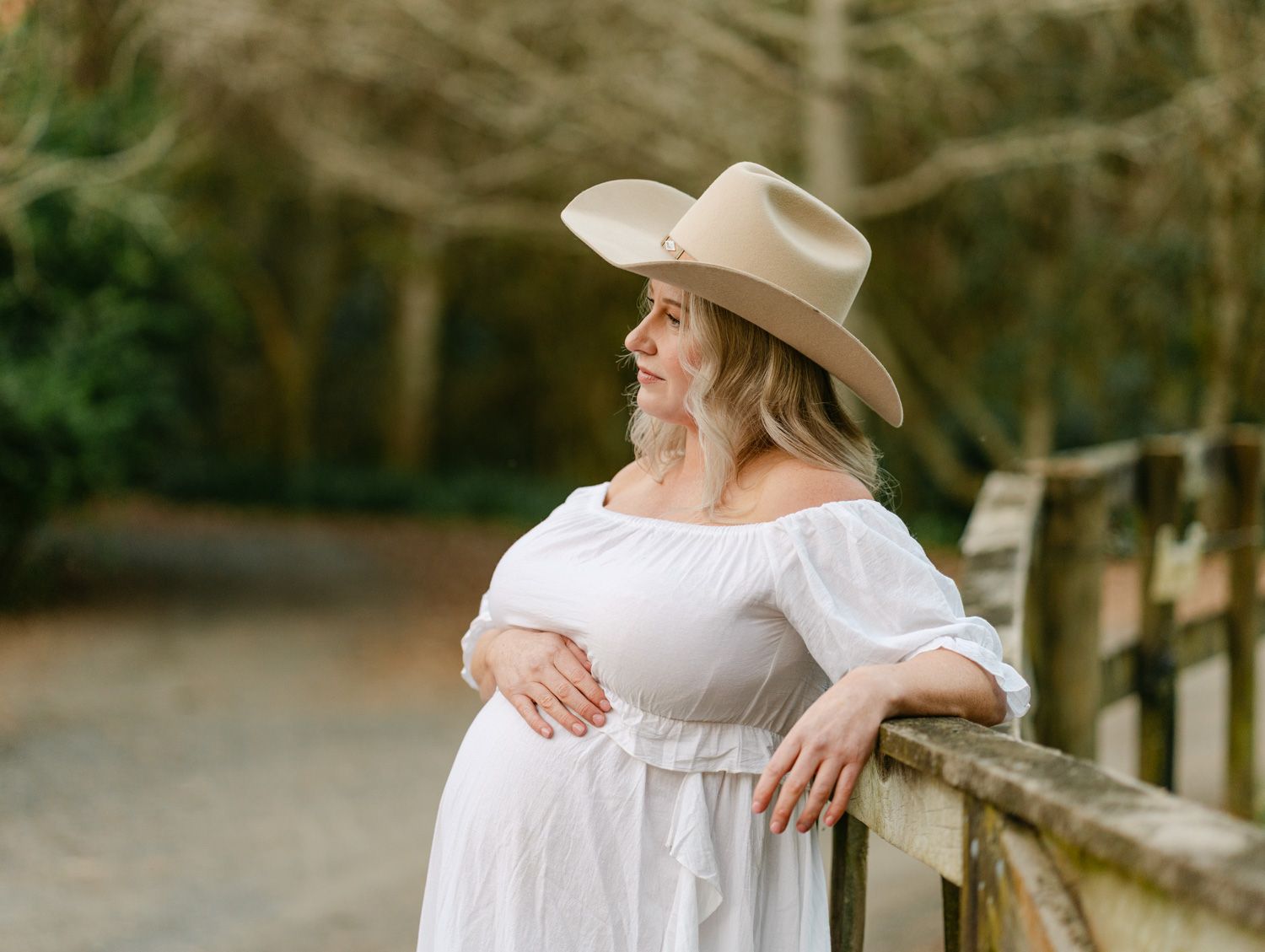 Pregnant woman in white off-shoulder dress and tan cowboy hat leaning on wooden fence in outdoor setting.