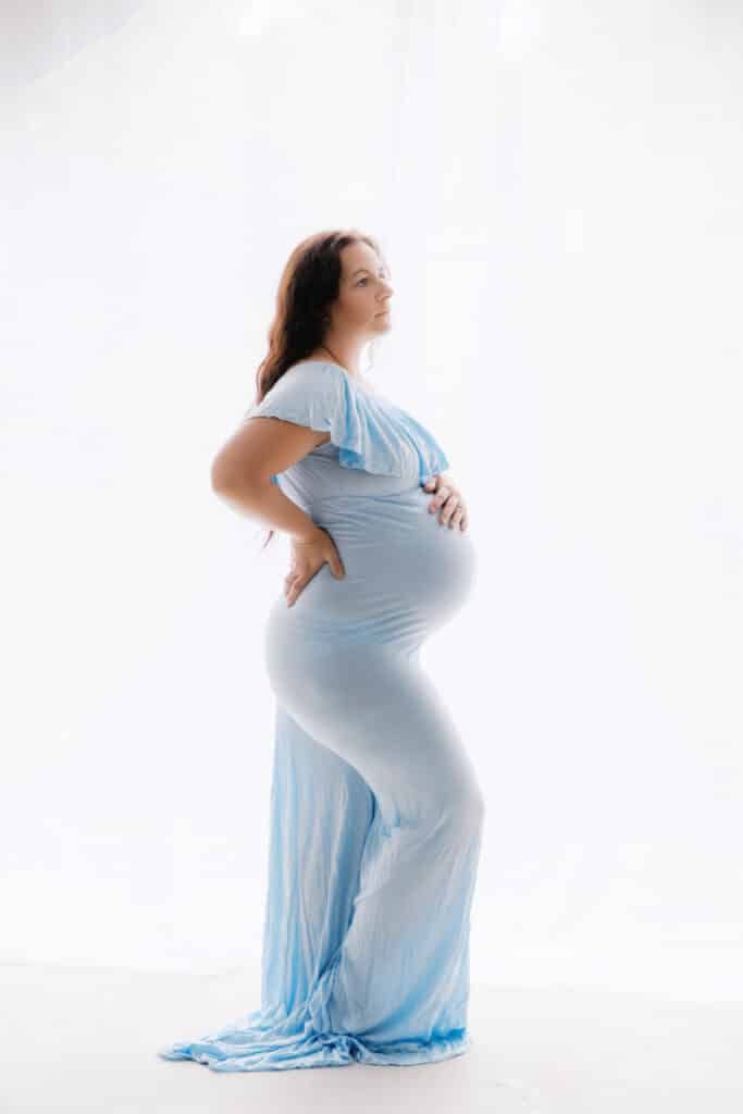 Pregnant woman in light blue maternity gown posing sideways, cradling baby bump in professional studio photo.