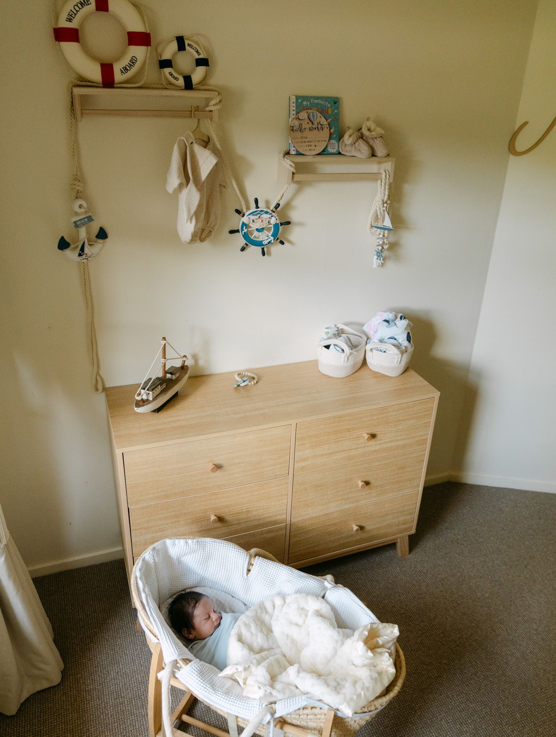 Newborn baby sleeping in a Moses basket beneath a nautical-themed nursery dresser with wall decor.