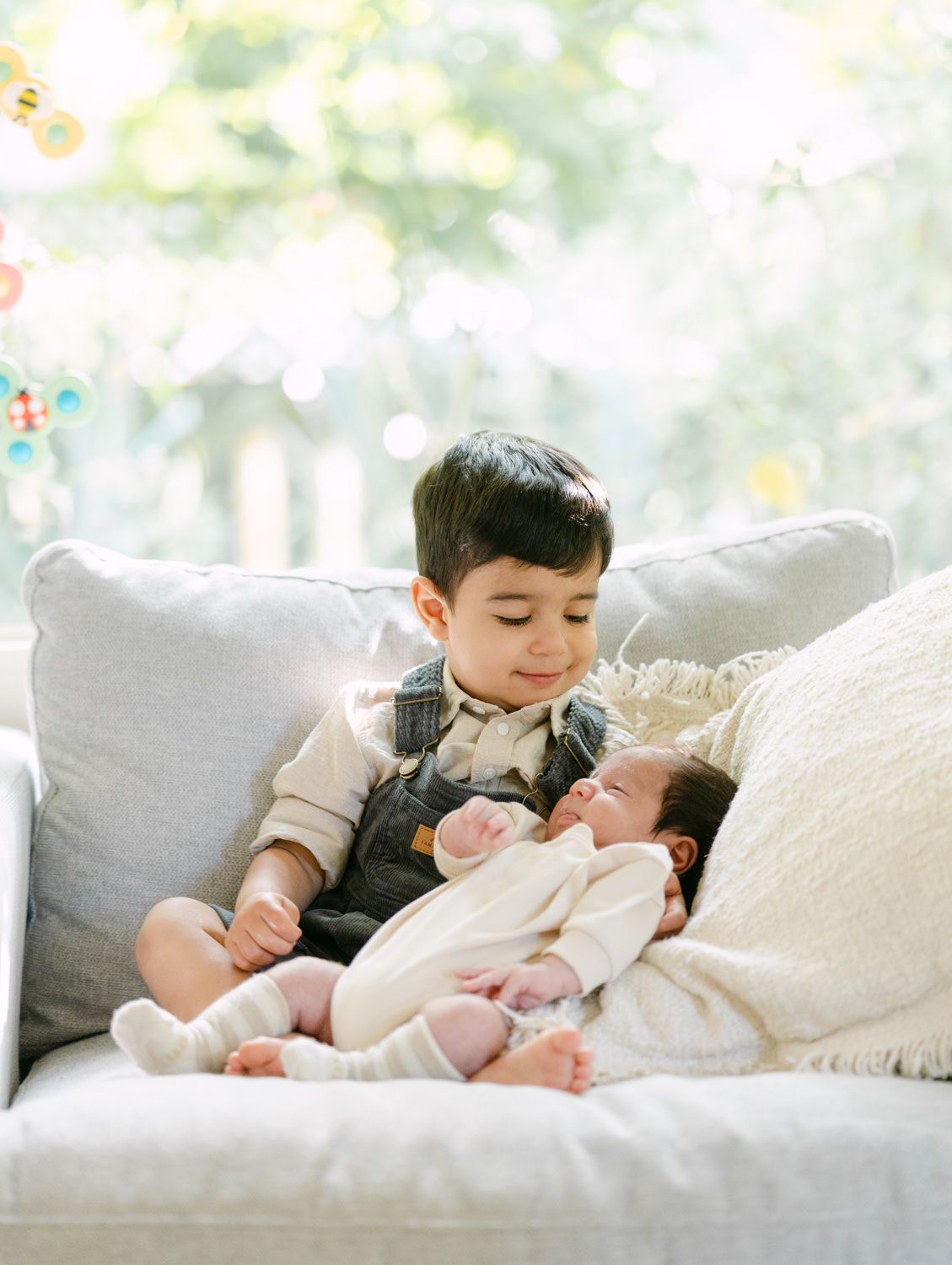 A young boy sits on a sofa smiling at the newborn baby resting in his lap near a window.