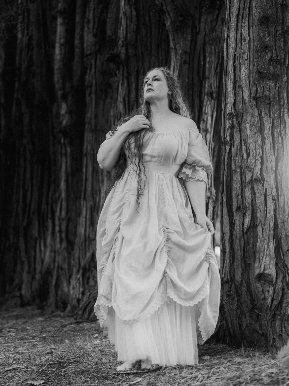 Black and white photo of a woman in a Victorian gown leaning against a large tree trunk in a forest.