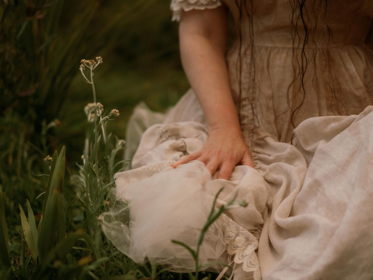 A young girl in a vintage white lace dress sits among wildflowers, gently holding fabric in a dreamy outdoor setting.