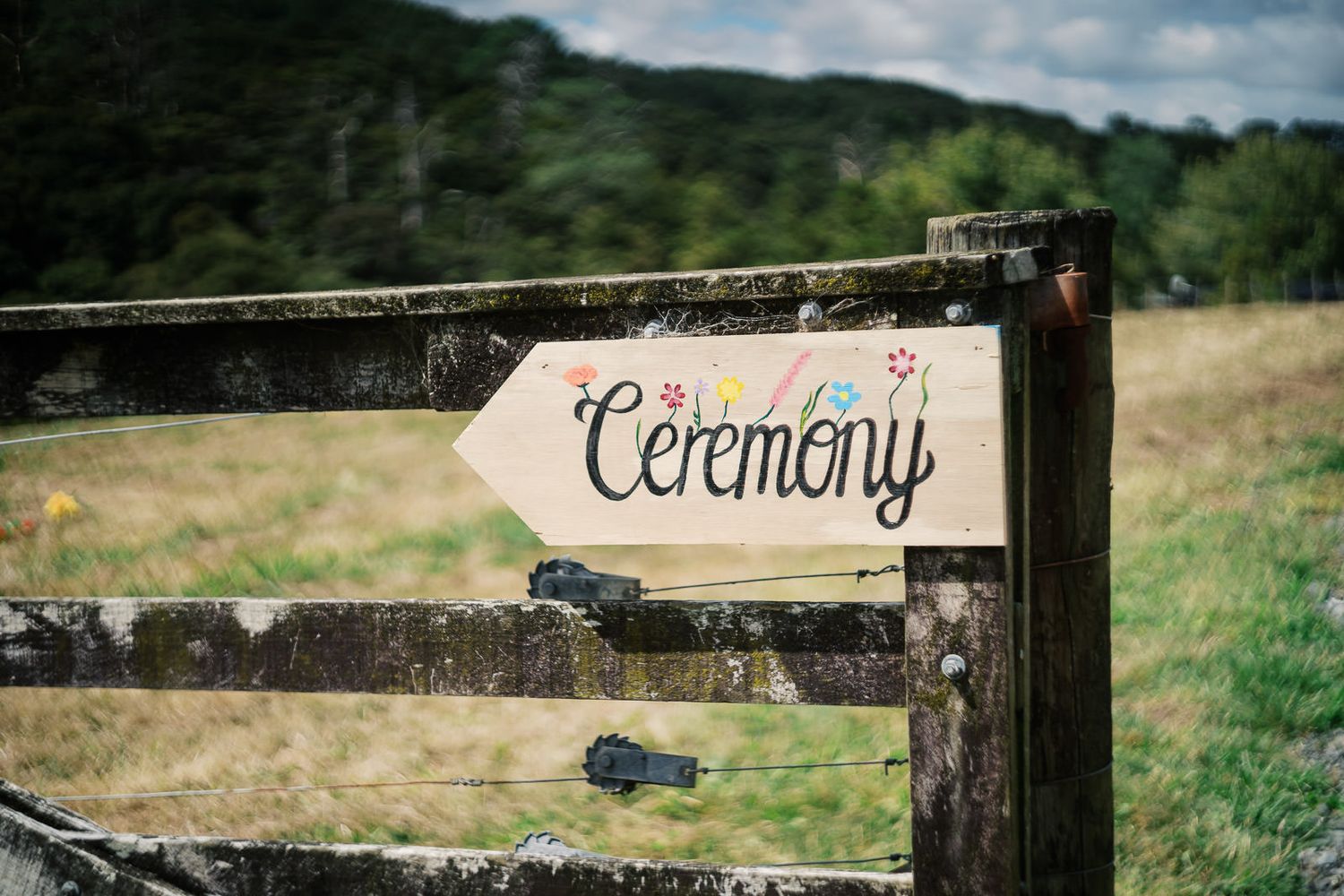 Rustic wooden arrow sign reading Ceremony with colorful floral details, mounted on a fence at an outdoor wedding venue.