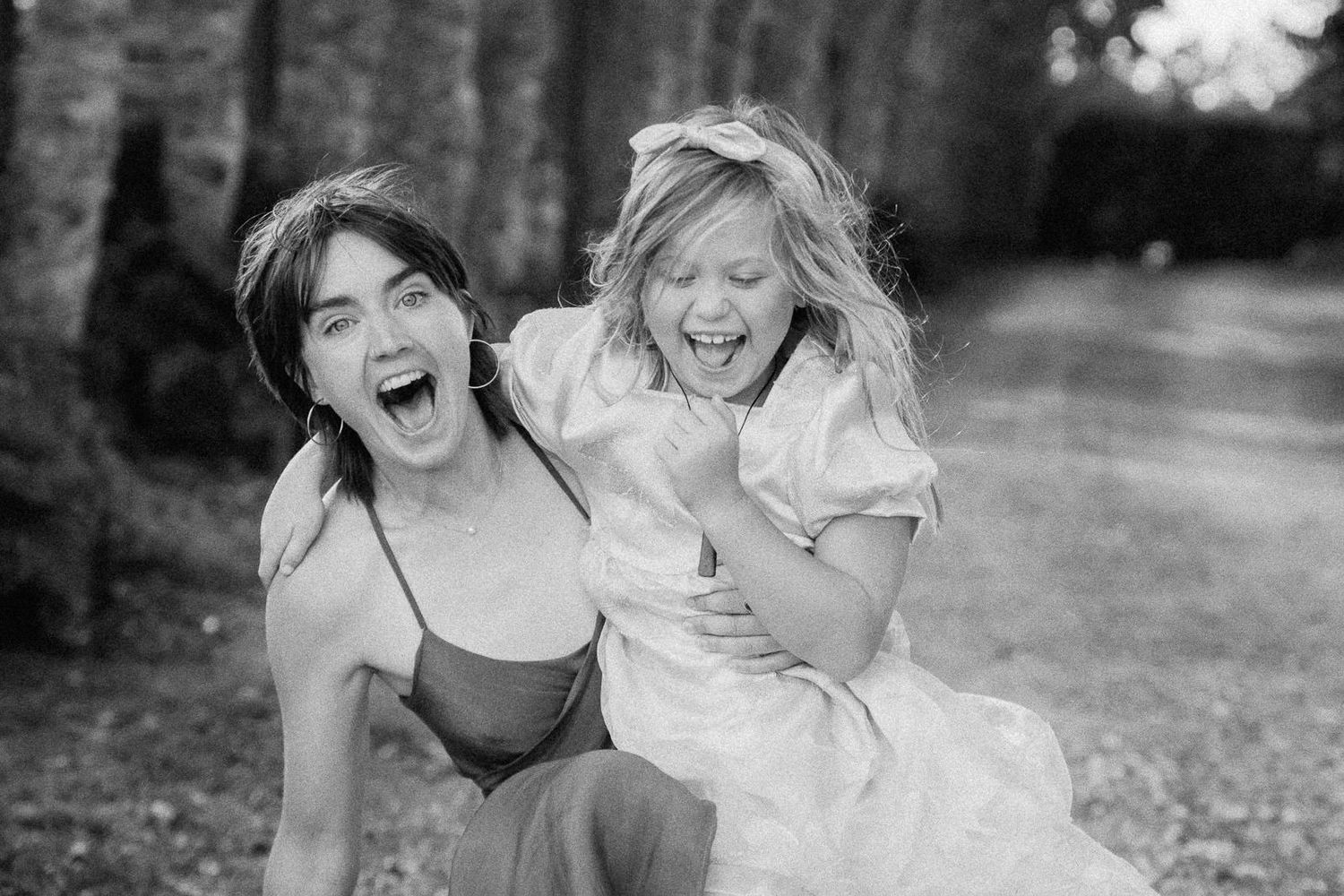 Two laughing girls share a joyful moment outdoors, one in a white dress, captured in black and white.