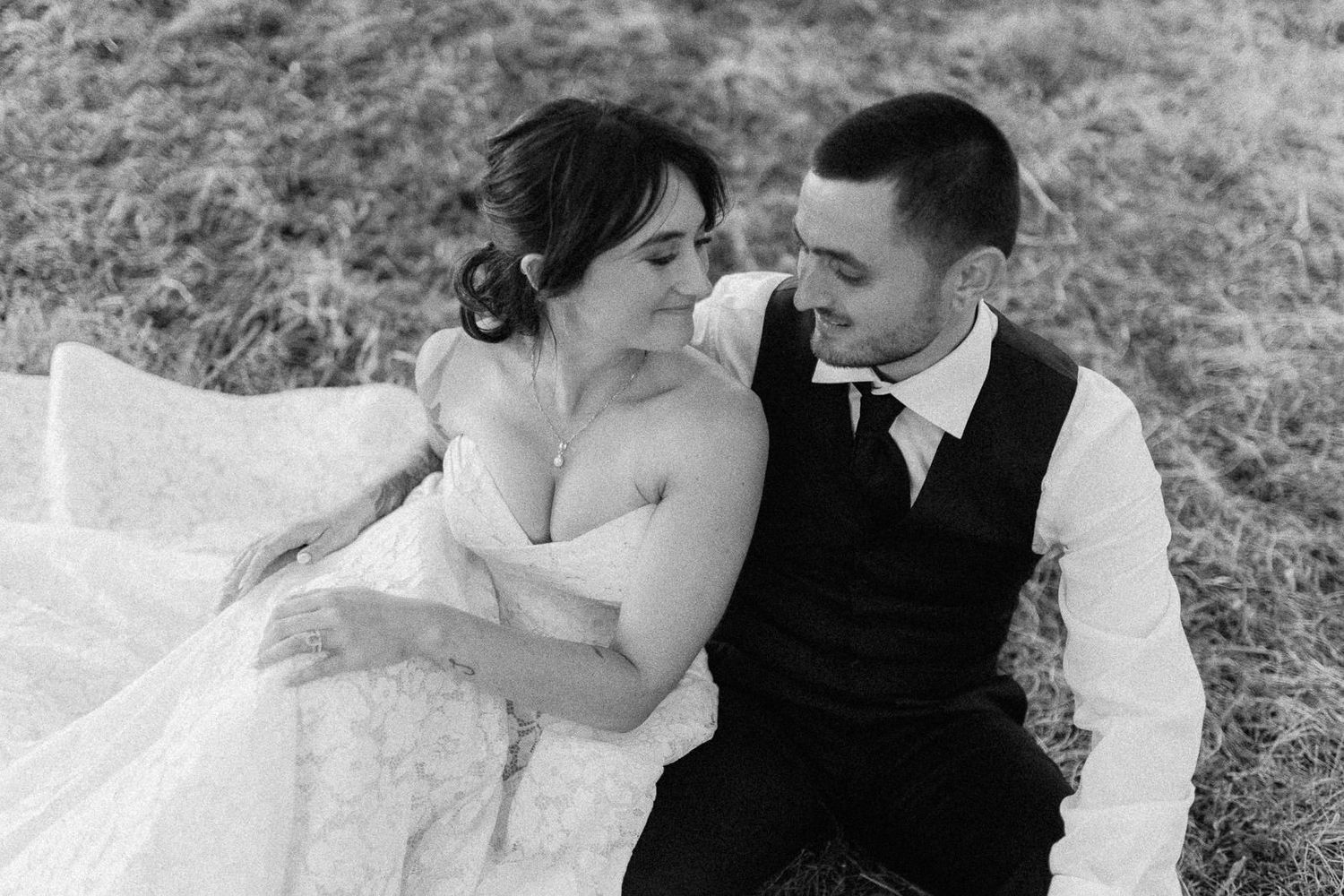Black and white wedding photo of bride and groom sitting together in a field, sharing an intimate moment.
