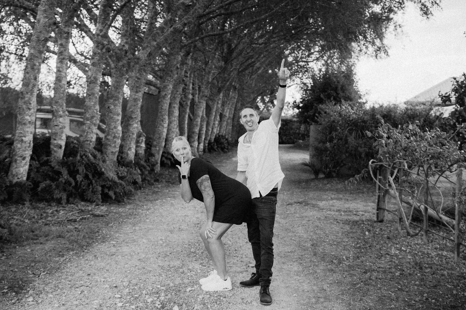 Black and white photo of a couple playfully posing on a tree-lined gravel path outdoors.
