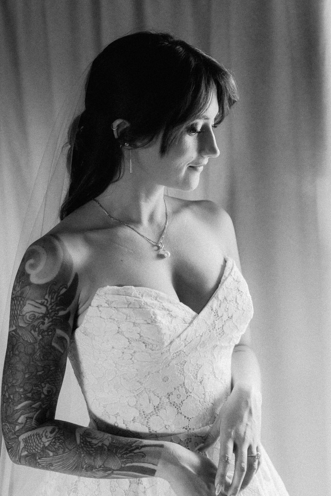 Black and white portrait of a tattooed bride in a strapless lace wedding gown with veil, looking elegantly to the side.
