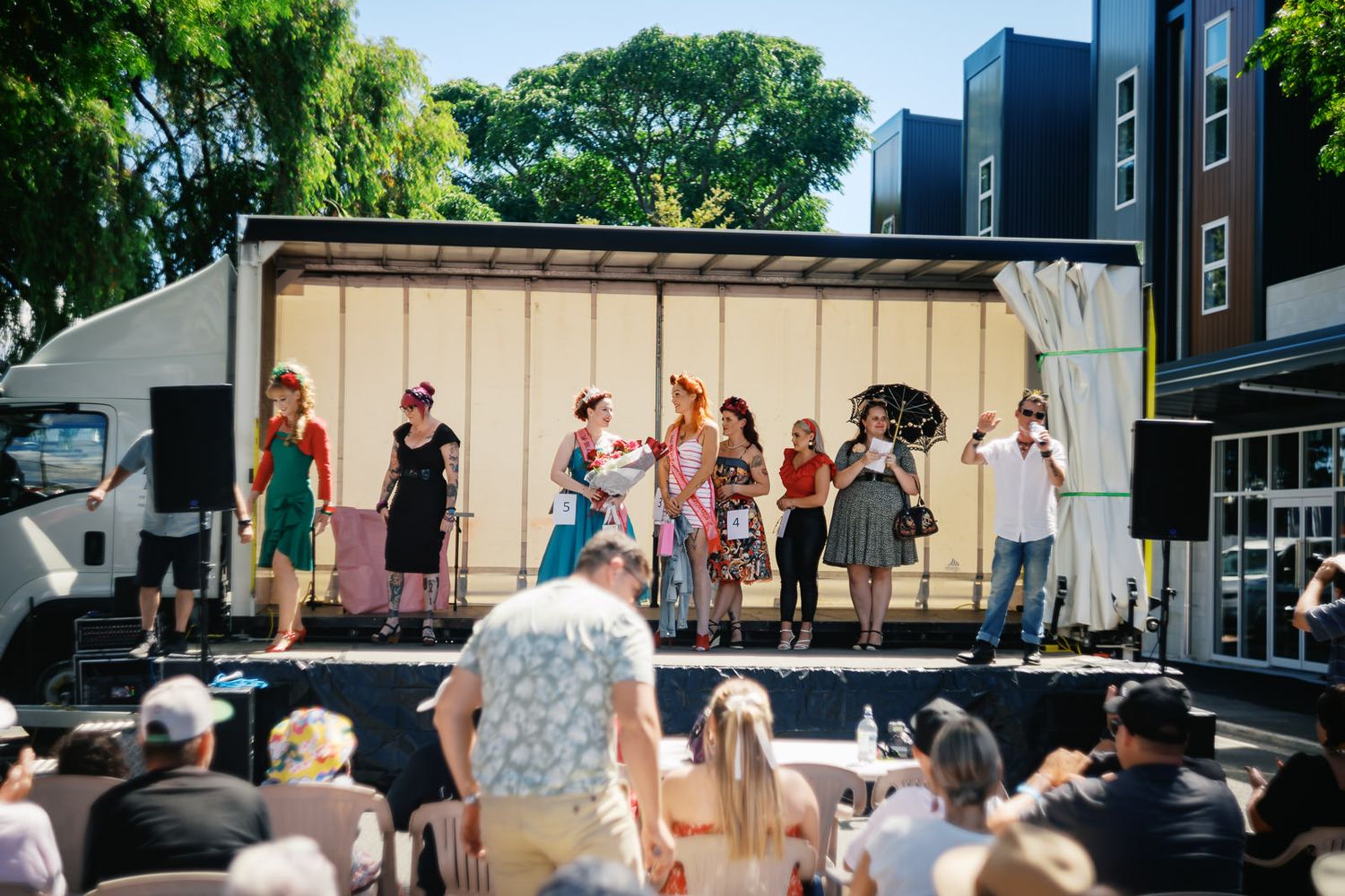 Outdoor performance on a truck stage with performers in colorful outfits entertaining a seated crowd on a sunny day.