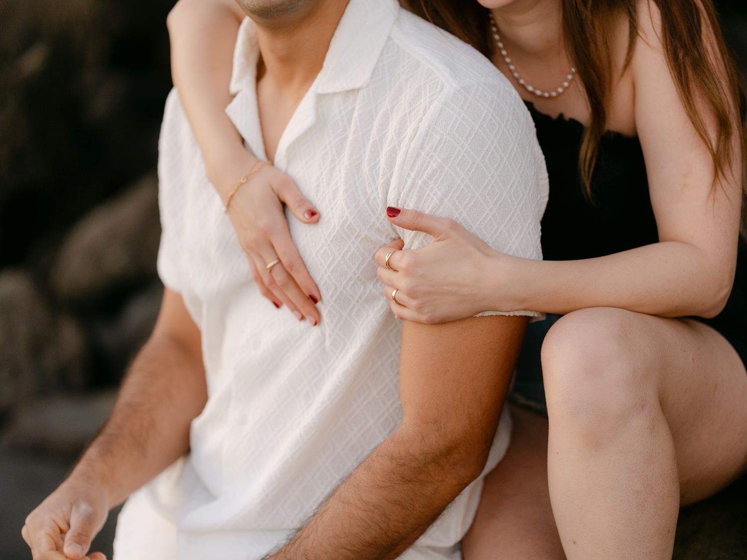 Couple sitting together outdoors, woman in black dress embracing man in white shirt, engagement photo session.