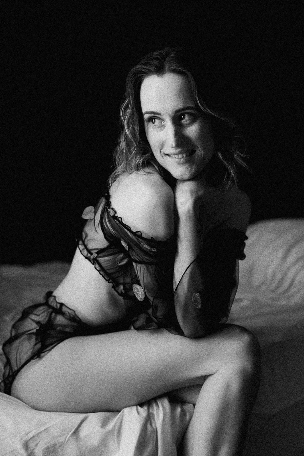 Smiling woman in sheer black lingerie poses on white bed in black and white boudoir photo.