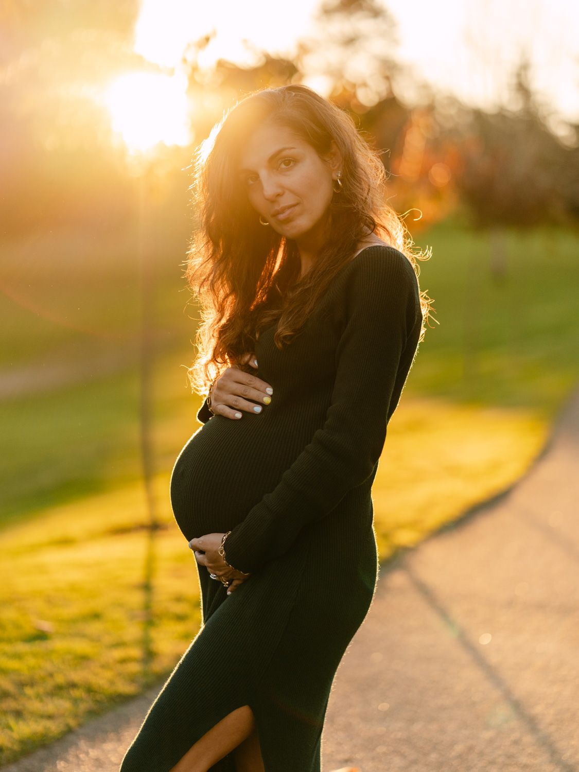 Pregnant woman in black dress holding baby bump at golden hour sunset in a park.