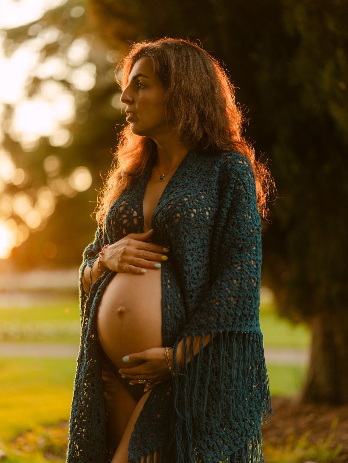 Pregnant woman in teal crochet wrap cradling baby bump at golden hour in a lush green park.