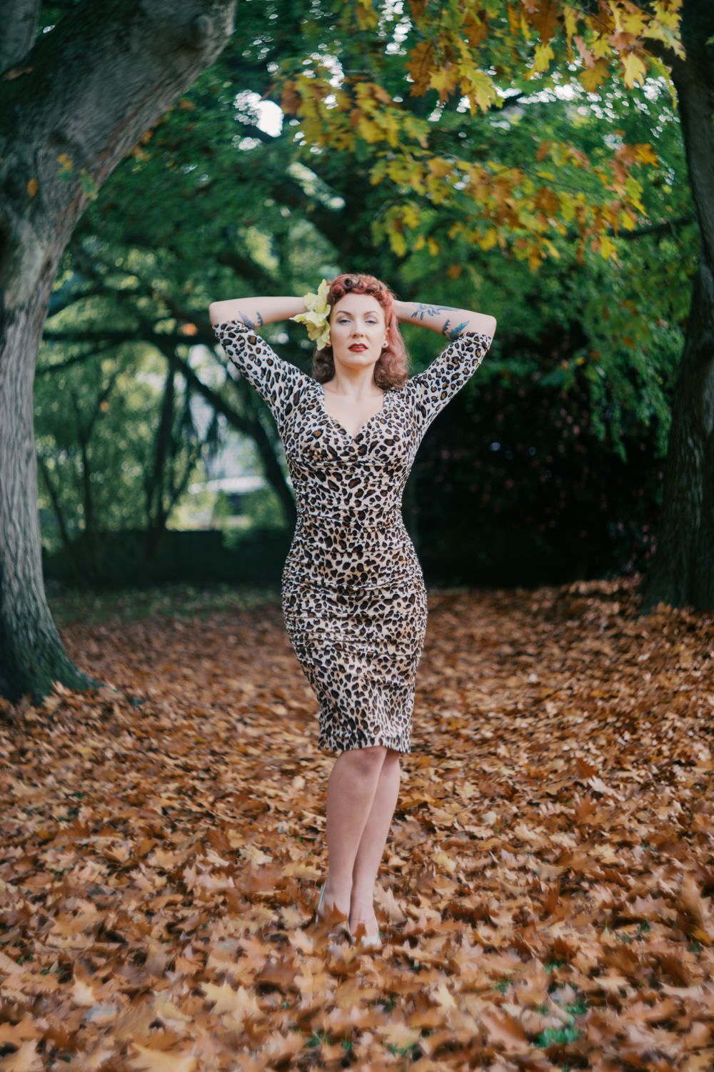 Woman in leopard print dress poses among autumn leaves in a park setting.