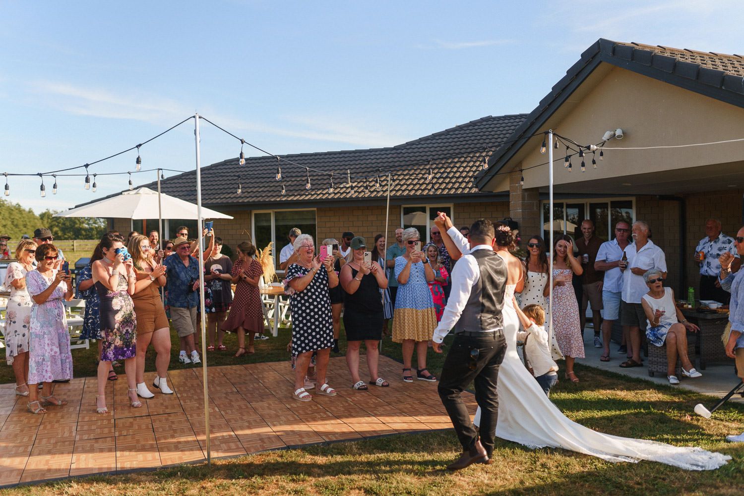 Bride and groom share first dance at outdoor backyard wedding reception with guests applauding around them.
