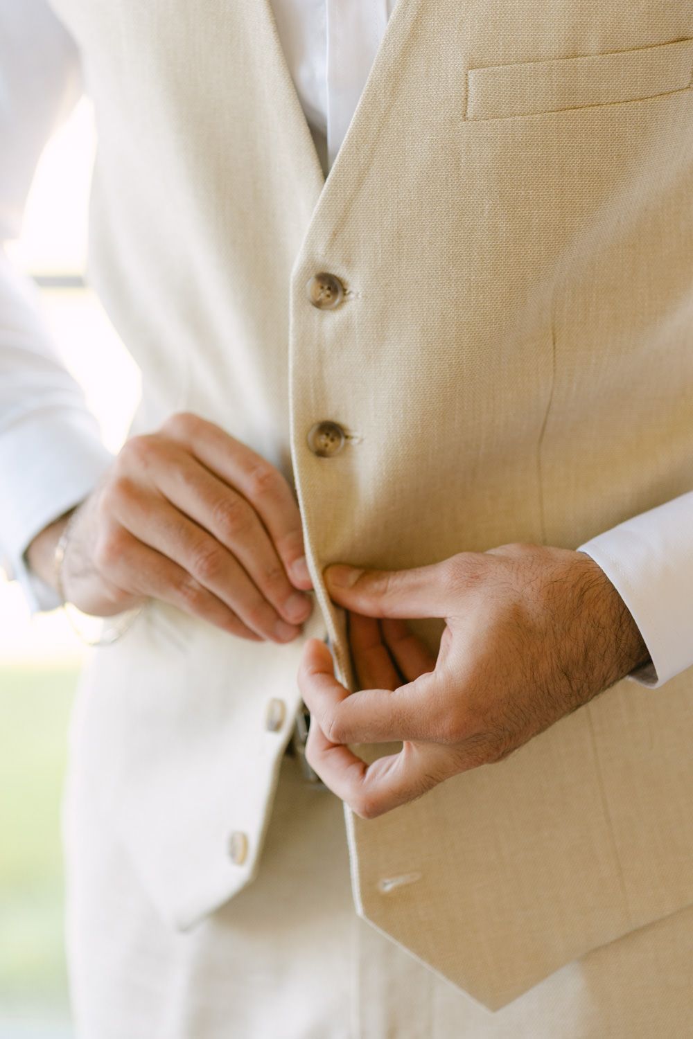 Man buttoning a beige linen vest suit jacket, preparing for a formal wedding ceremony.