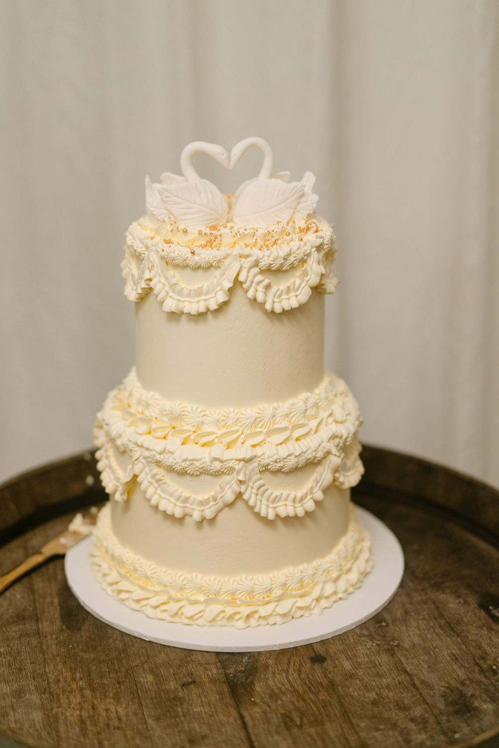 Elegant three-tier white wedding cake with ruffled frosting and swan topper on rustic wooden barrel.
