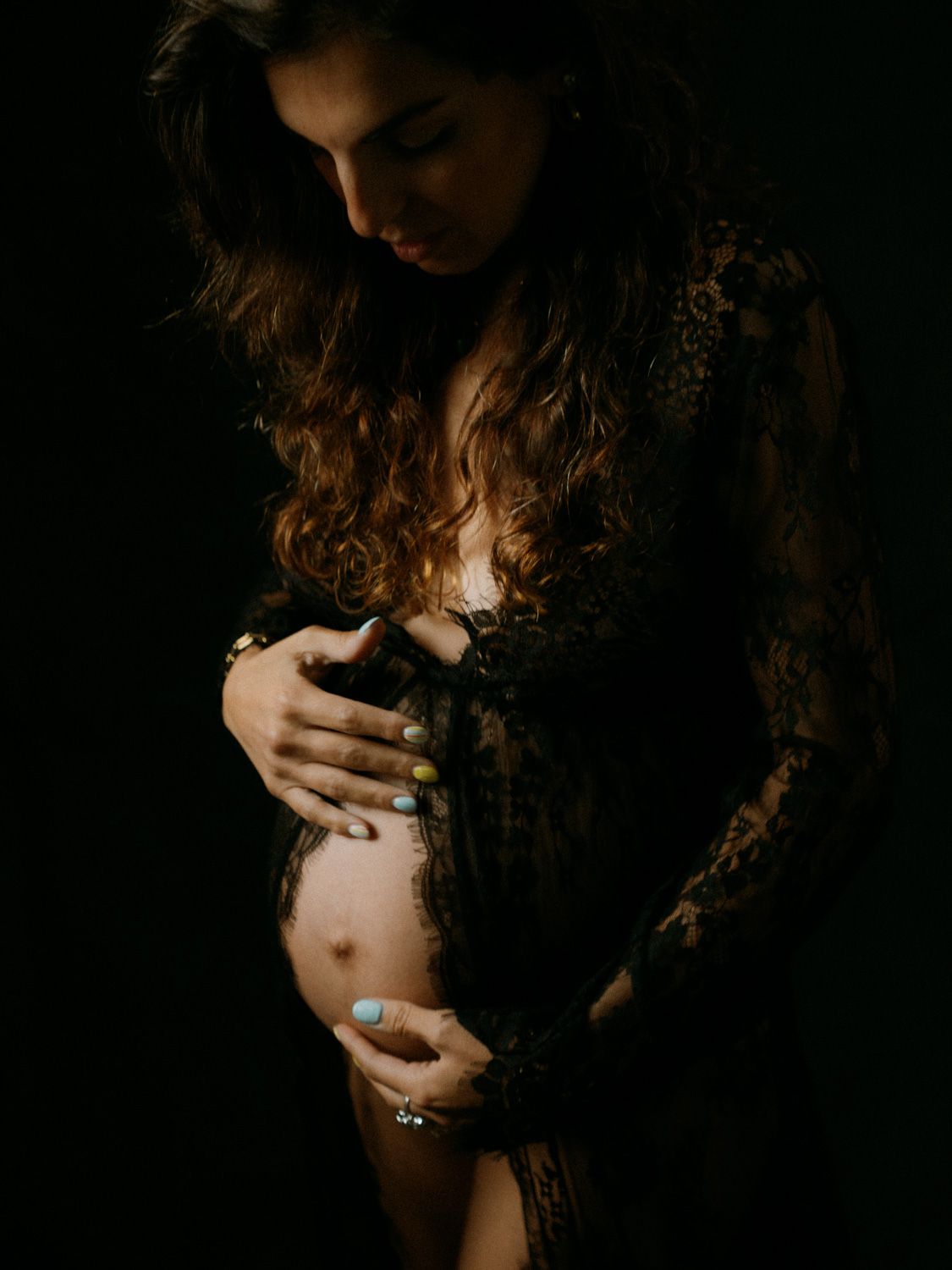 Pregnant woman in black lace dress cradling her baby bump against a dark background.