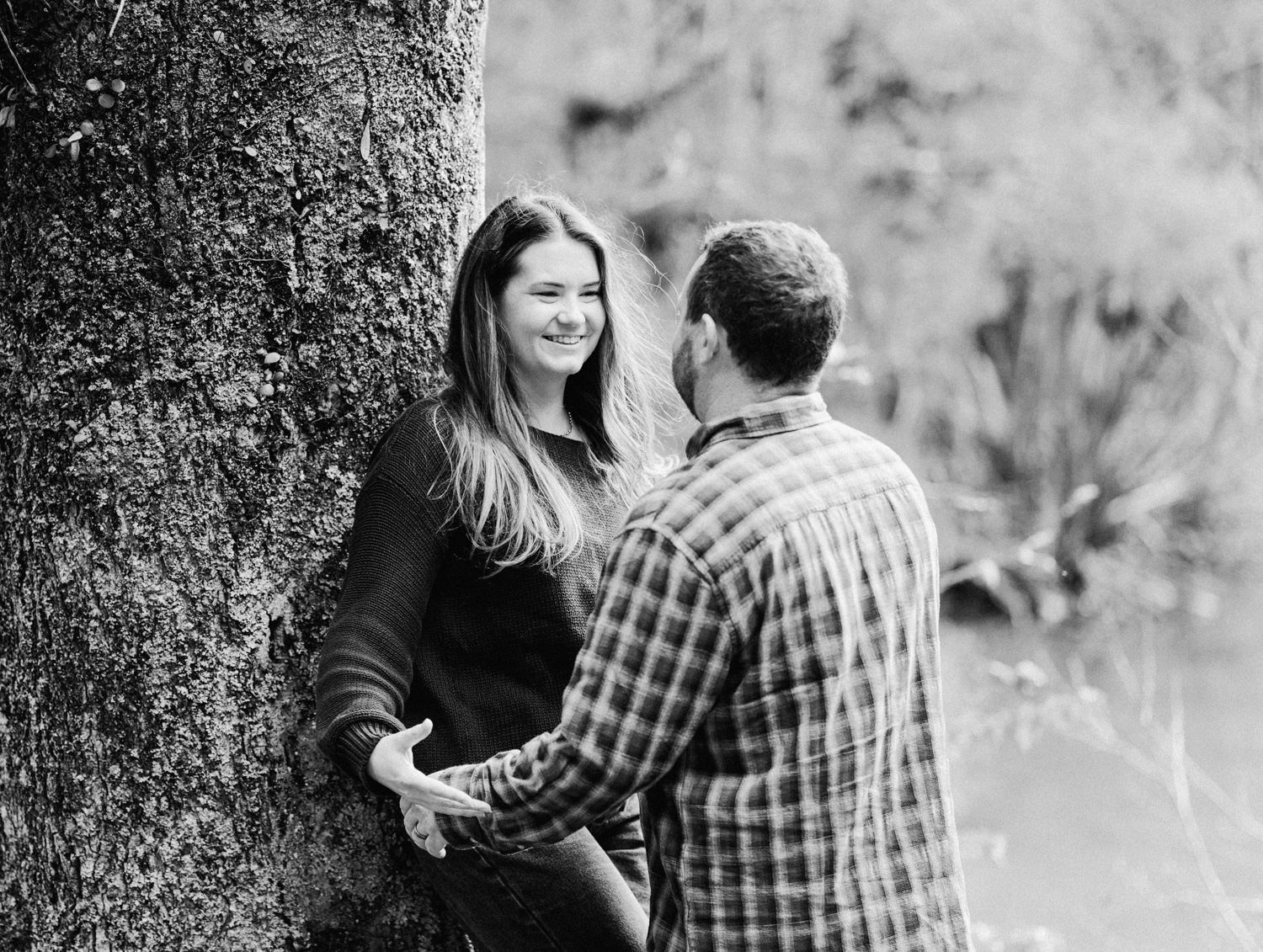 Black and white photo of a couple holding hands and smiling near a large tree outdoors.