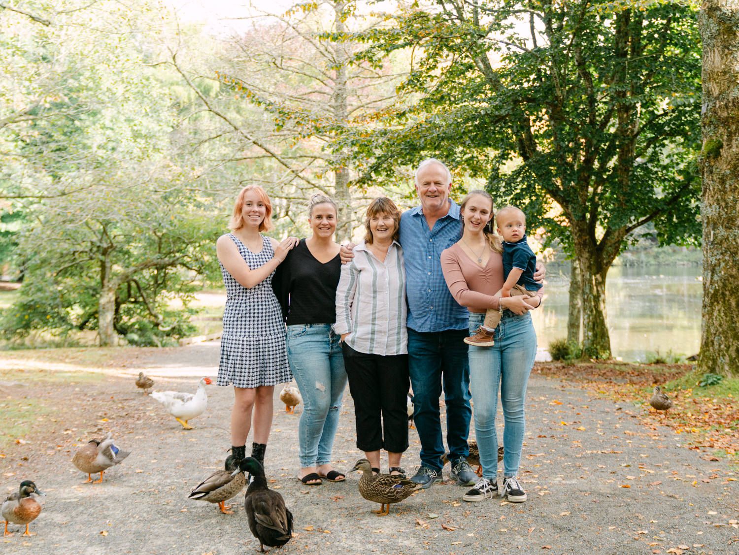 Family of six posing outdoors near a pond with ducks, surrounded by lush green trees in a park setting.