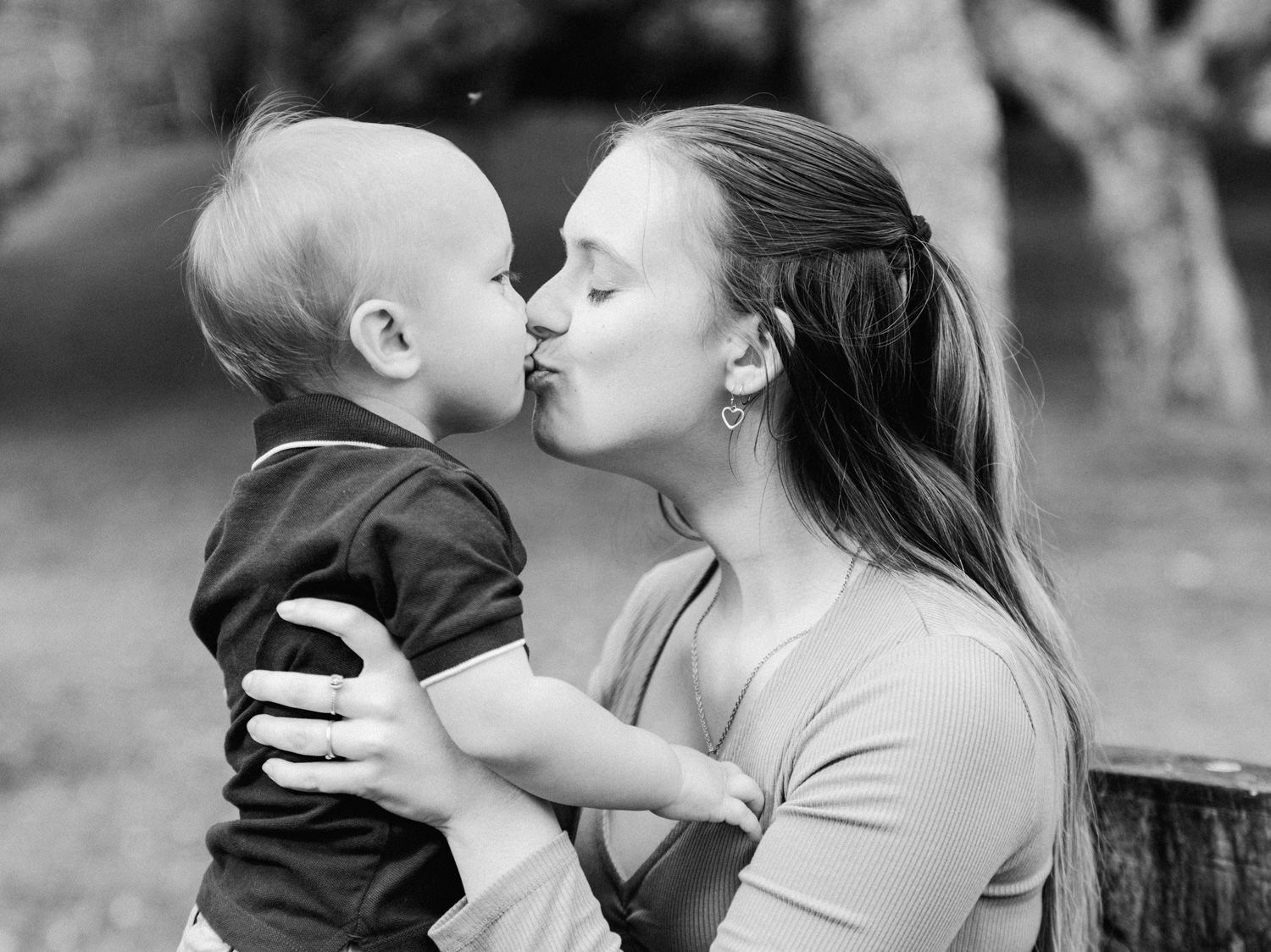 Black and white photo of a young mother kissing her baby boy outdoors in a park setting.