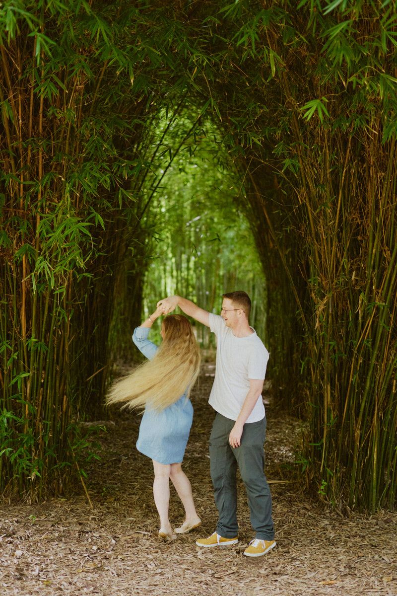 Couple dancing together in a lush bamboo forest tunnel, woman's hair flowing as man twirls her on a sunlit path.