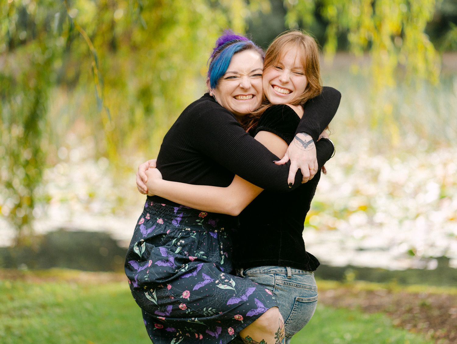Two women hugging and smiling outdoors near a willow tree, one with blue hair wearing a floral skirt.