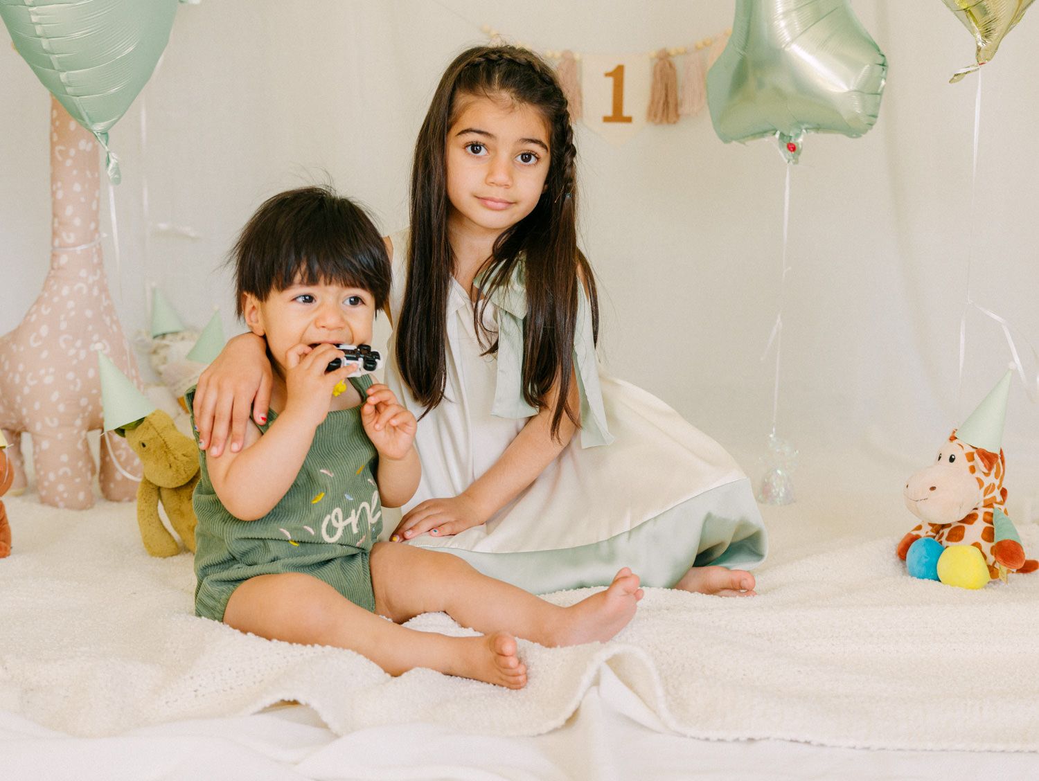 Two young children pose together on a white bed surrounded by sage green balloons and stuffed animals at a birthday party.