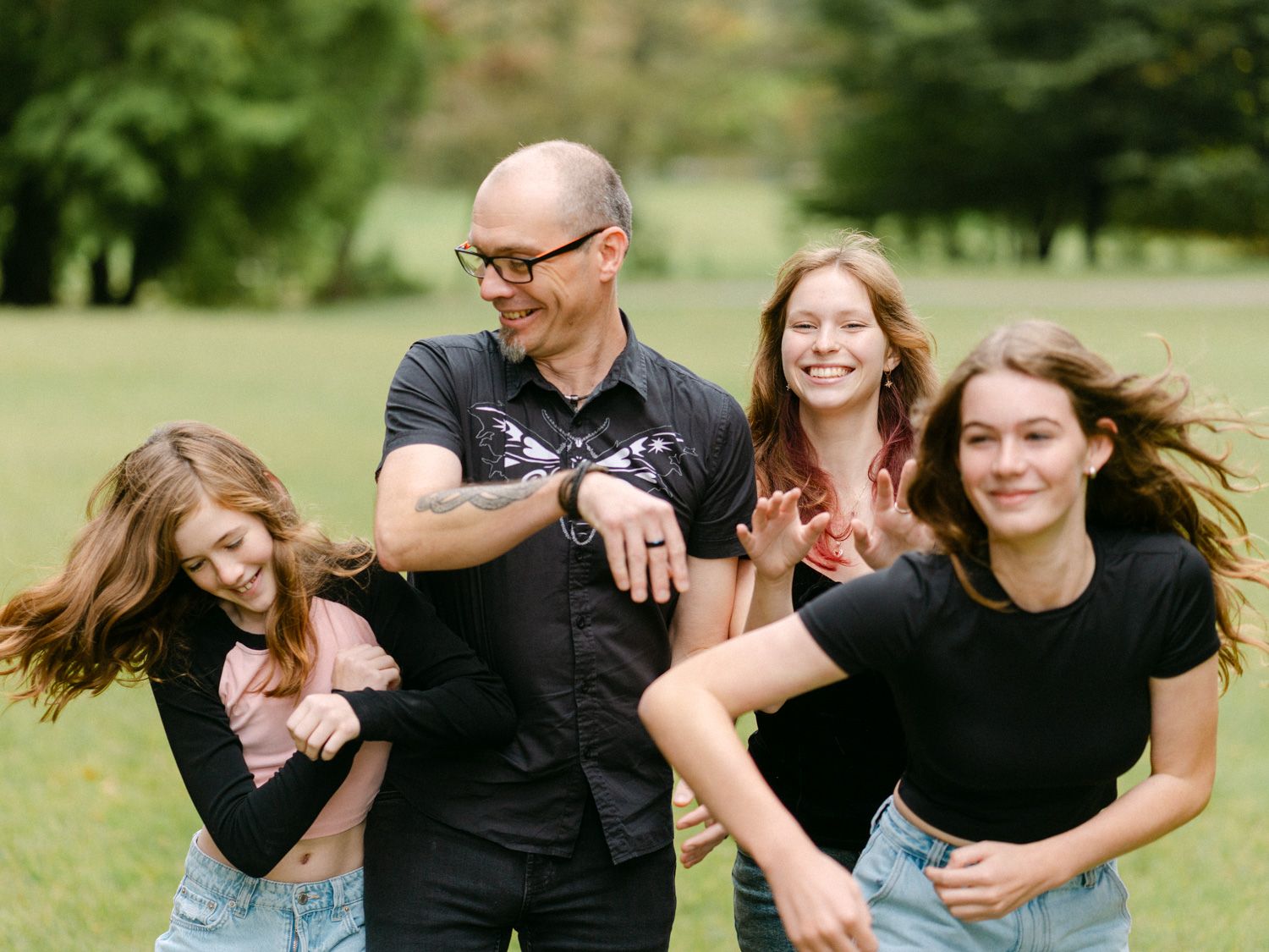 A man and three young women laugh and dance together outdoors in a lush green park during a family photo session.