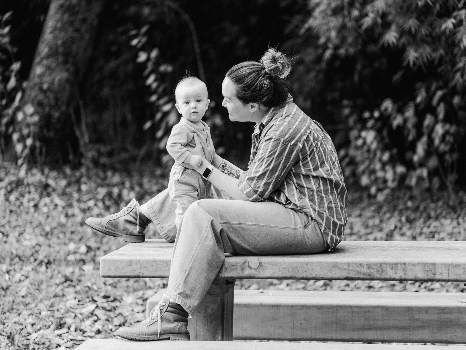 Black and white photo of a mother sitting on a bench with her baby in a peaceful outdoor park setting.