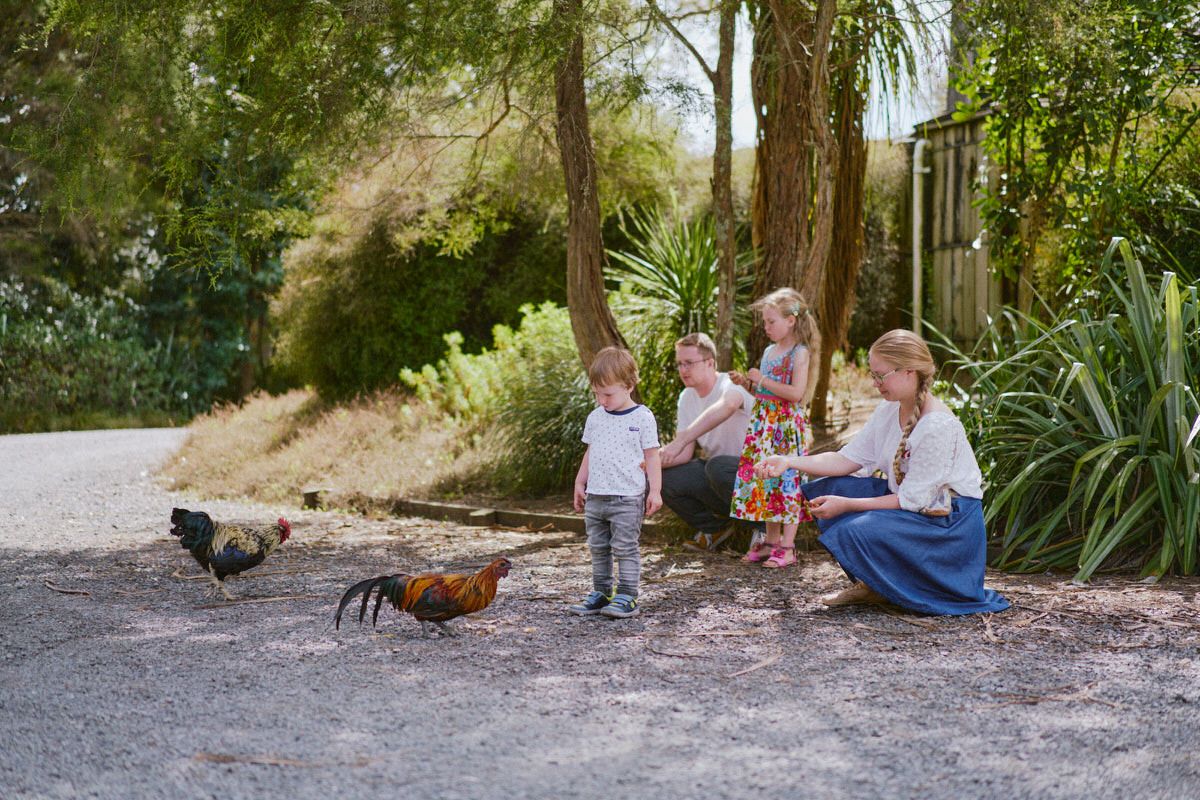 Family with children feeding chickens outdoors on a gravel path surrounded by lush green trees and garden.