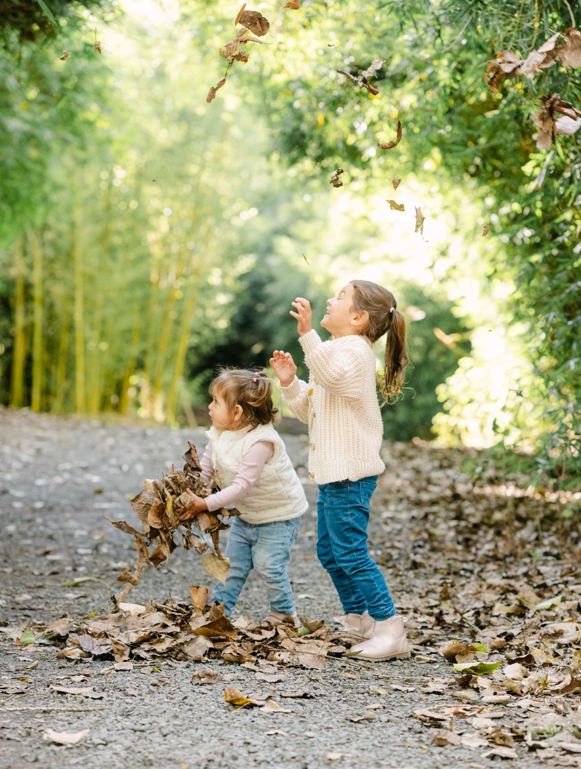 Two young girls in cream sweaters and jeans joyfully toss autumn leaves on a tree-lined path.