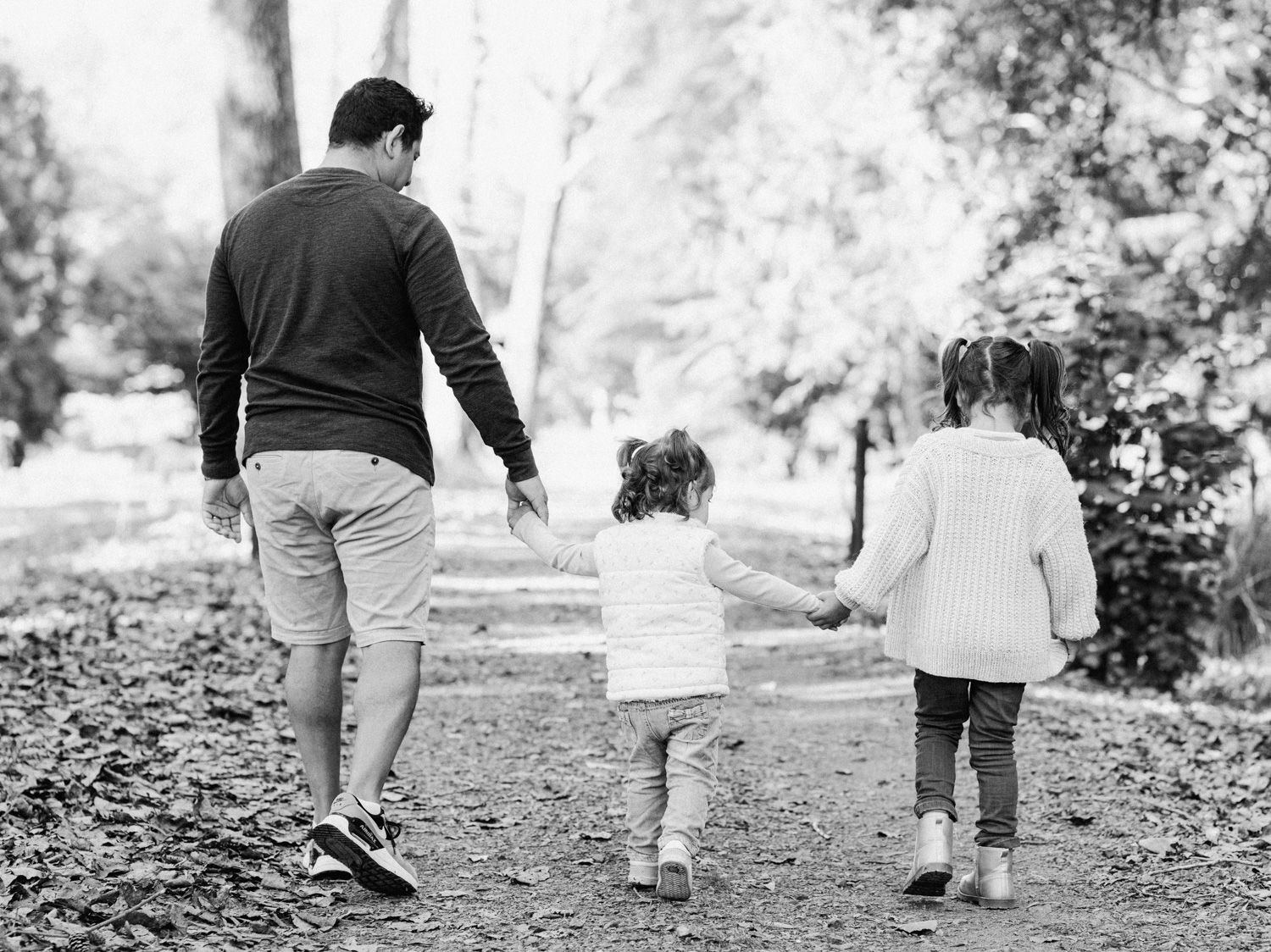 Black and white photo of a father walking hand in hand with two young daughters on a tree-lined path.