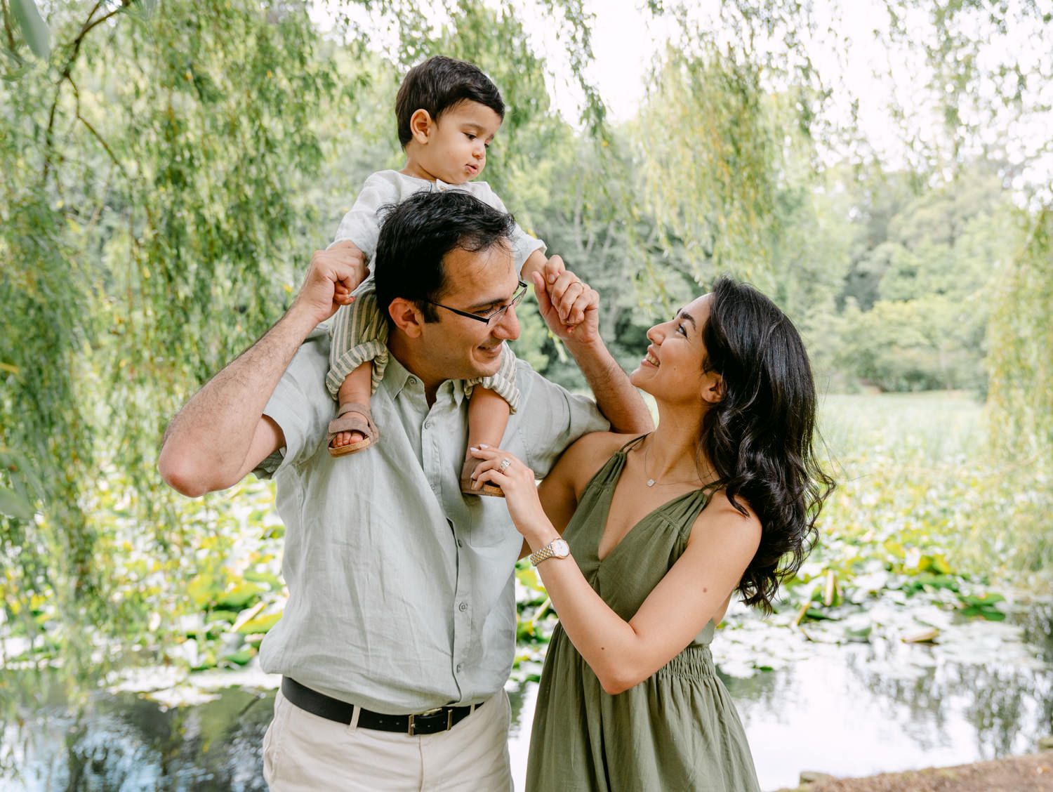 Happy family portrait outdoors, father carrying toddler on shoulders while mother smiles, lush green garden pond backdrop.