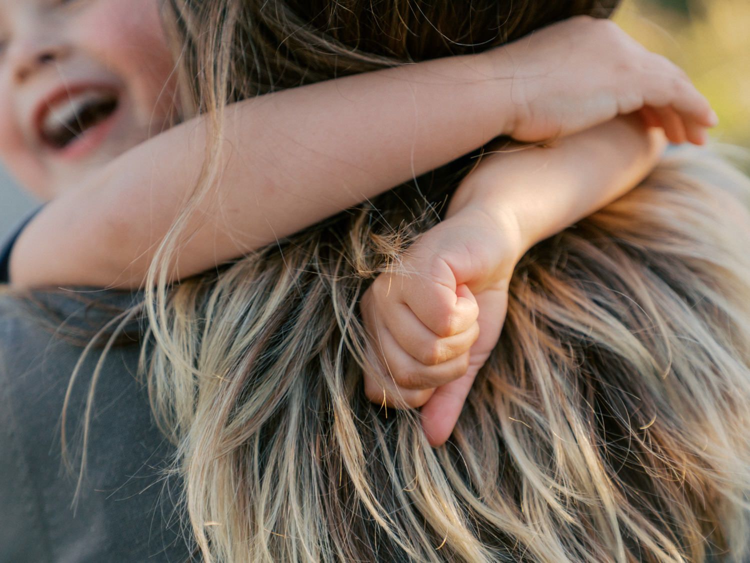 A laughing child hugs an adult with long blonde hair, showing a joyful and loving embrace from behind.