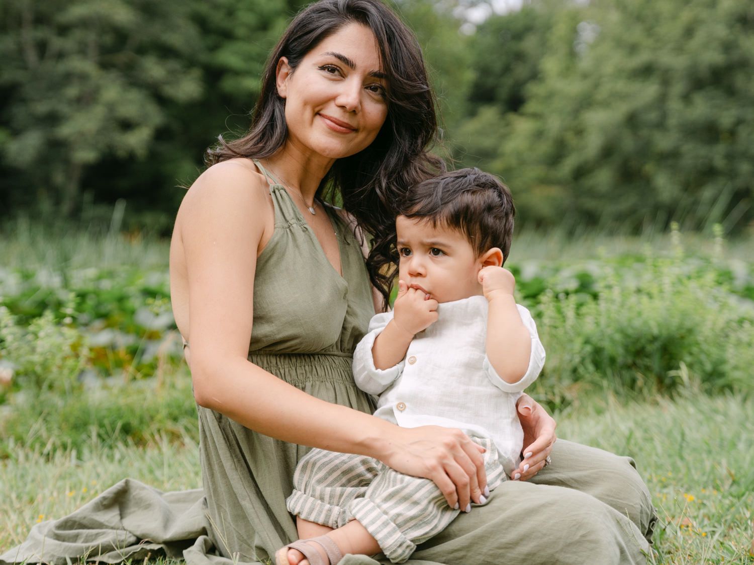 A mother in a green dress sits outdoors holding her baby in a lush garden setting.