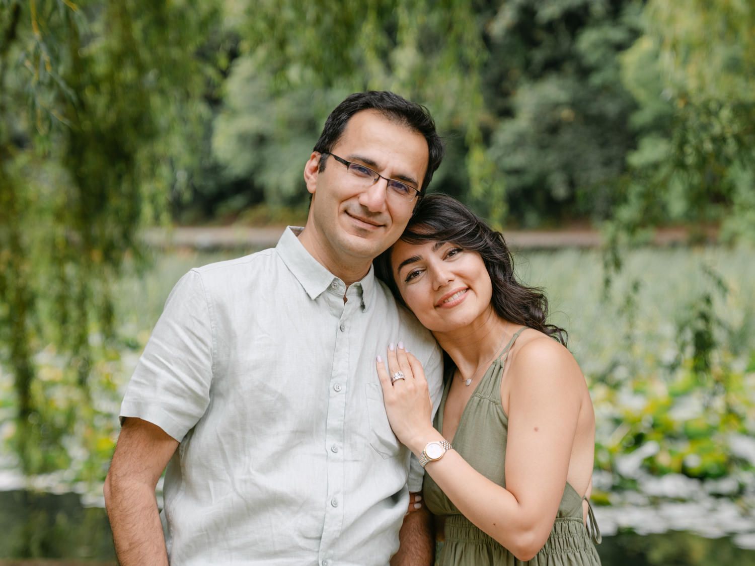 Smiling couple poses outdoors near a weeping willow tree and pond in a lush green garden setting.