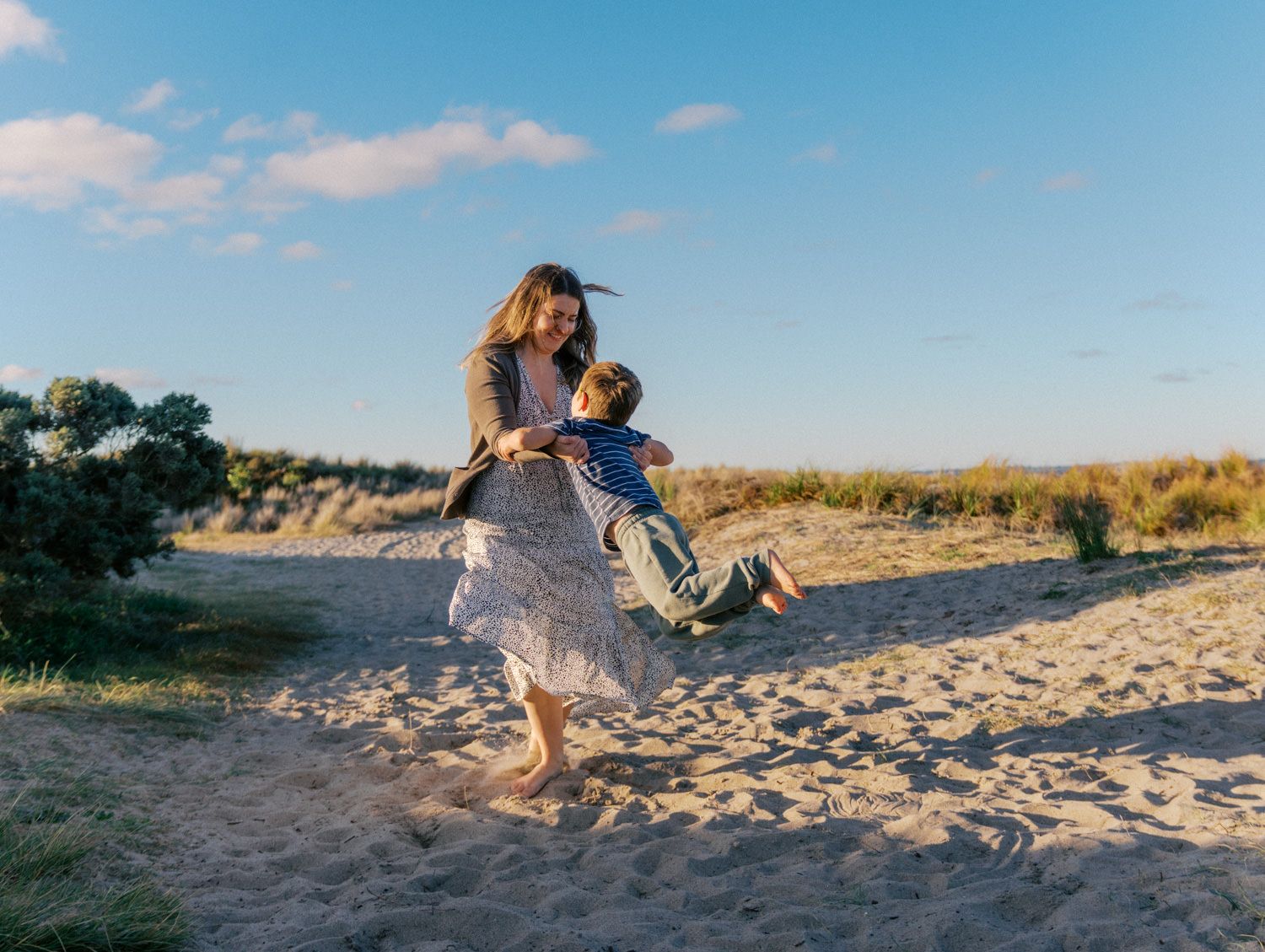 A joyful mother swings her young child on a sandy beach under a bright blue sky.