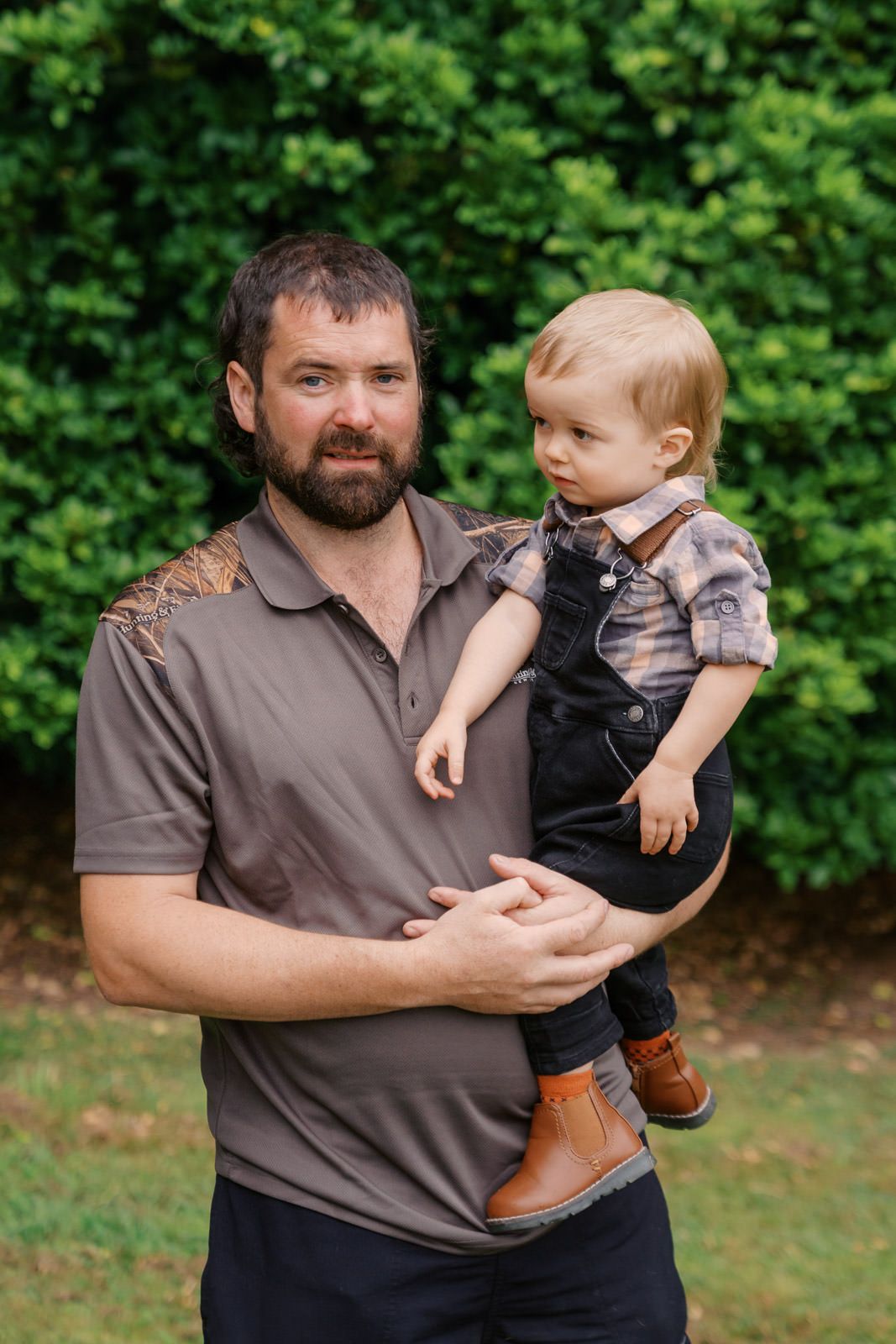 Bearded father holding toddler boy in overalls outdoors with green bushes background.
